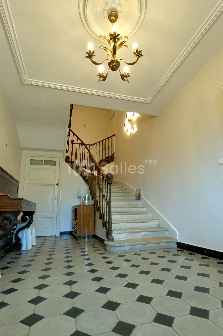 Escalier en bois avec rampe en fer forgé dans un hall lumineux au sol carrelé en motifs géométriques.