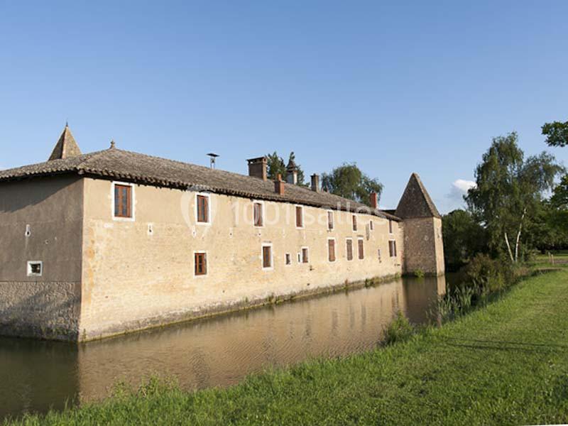Bâtiment en pierre avec toit en tuiles, entouré d’un fossé d’eau et bordé de végétation sous un ciel dégagé.