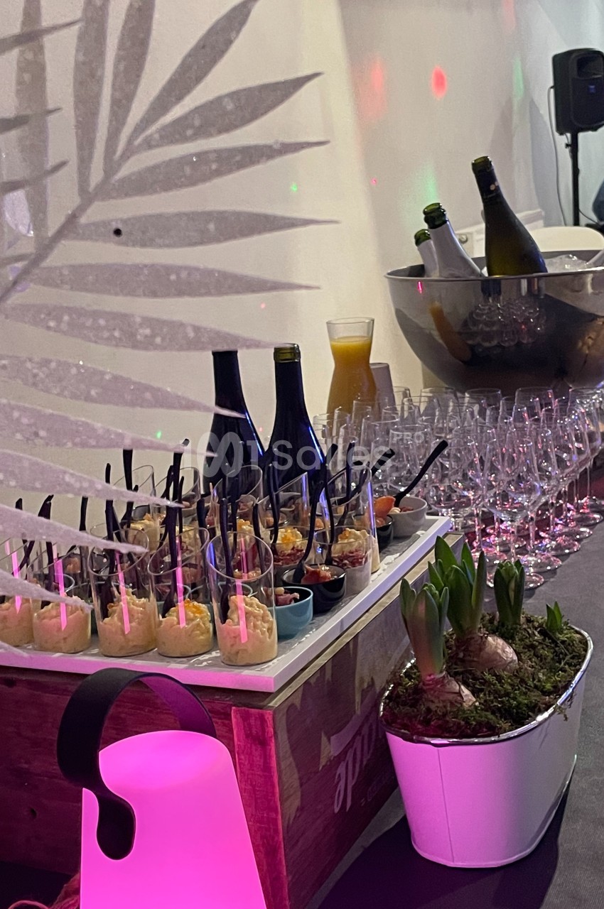 Buffet avec verrines, bouteilles de vin, jus d’orange, verres et décoration végétale sur une table éclairée.
