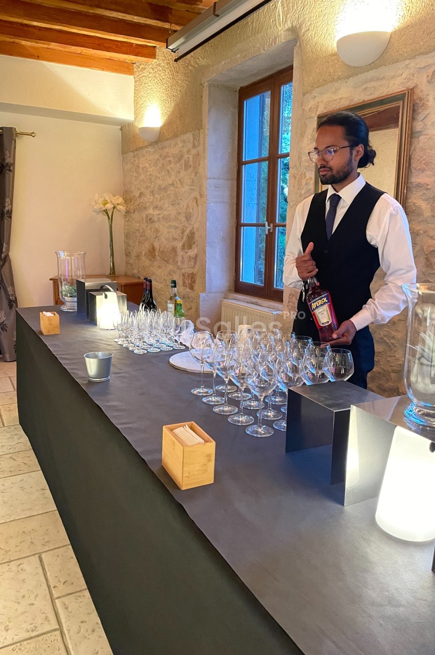 Un serveur en tenue formelle prépare un bar avec des verres à vin alignés dans une salle en pierre éclairée.