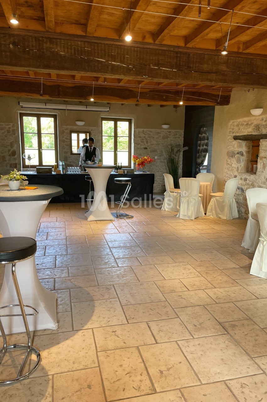Salle lumineuse avec tables hautes, chaises habillées de housses blanches, et un buffet en arrière-plan.