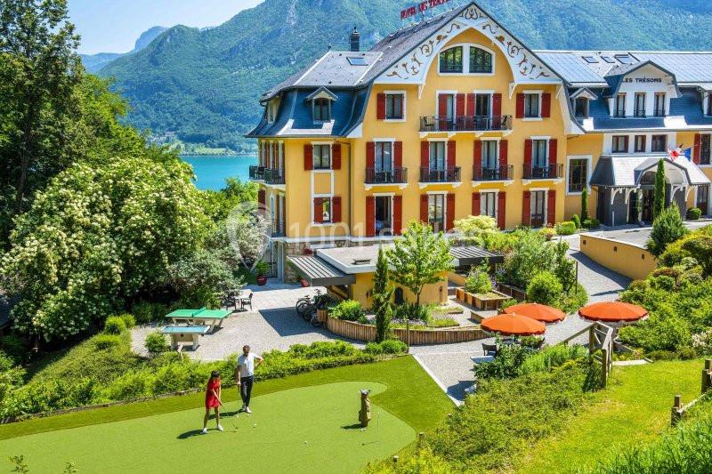 Un bâtiment jaune avec volets rouges, entouré de verdure, près d'un lac et des montagnes, avec un mini-golf au premier plan.