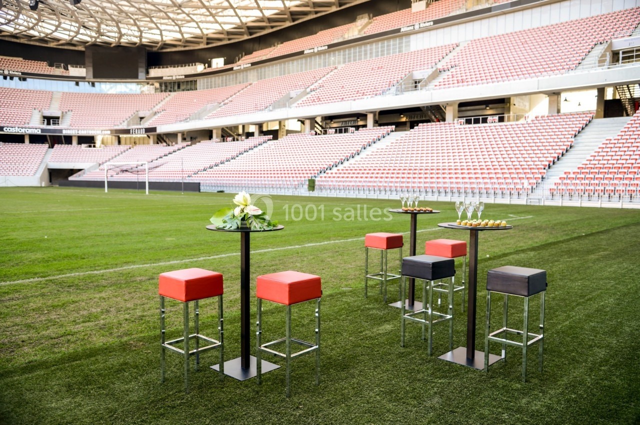Tables hautes avec tabourets colorés disposés sur une pelouse de stade, entourées de gradins vides.