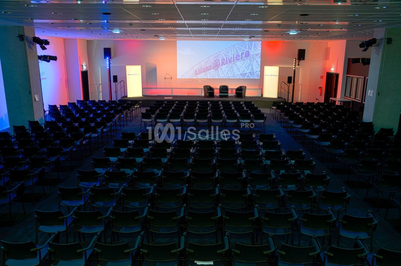 Salle de conférence vide avec des rangées de chaises noires, un écran de projection et un éclairage coloré.