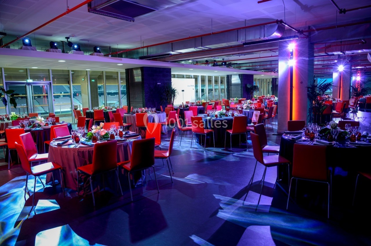 Salle de réception moderne avec tables rondes dressées, chaises rouges et éclairage coloré.