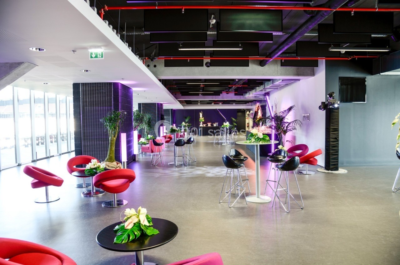 Salle moderne avec mobilier design, tables hautes, fauteuils rouges et plantes, éclairage coloré et ambiance lumineuse.