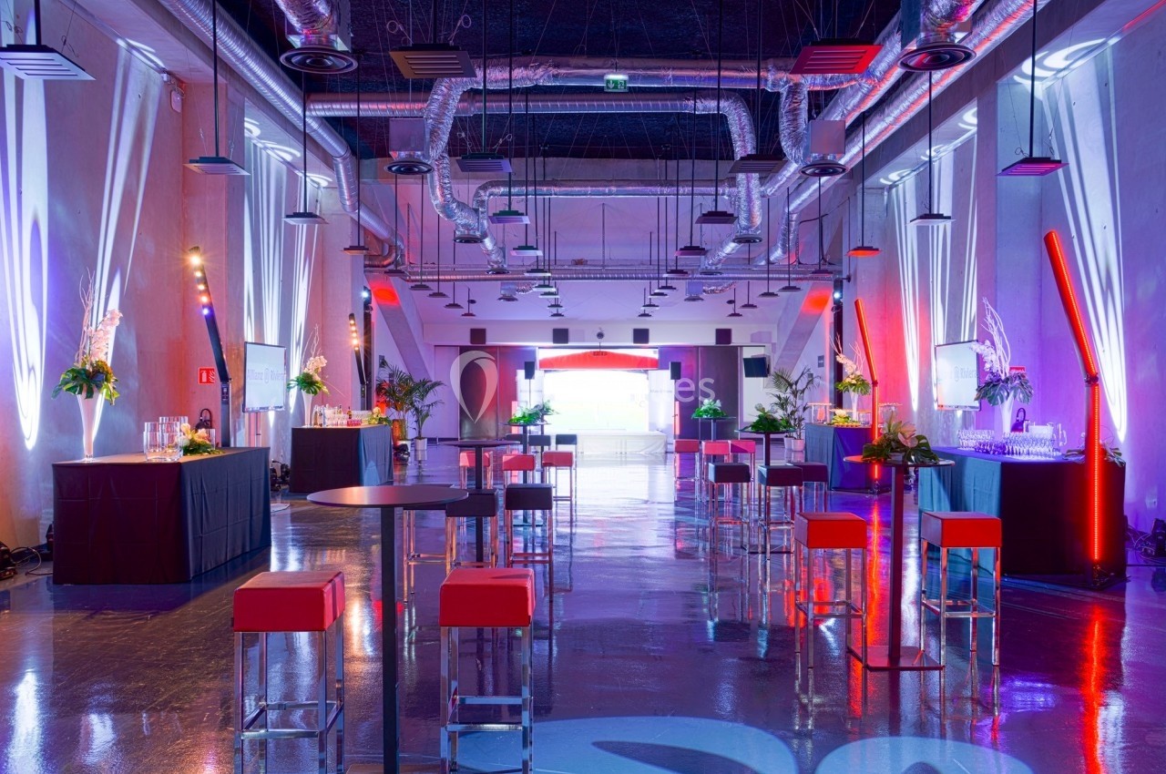 Salle moderne aménagée pour un événement, avec tables hautes, tabourets rouges et éclairage coloré.
