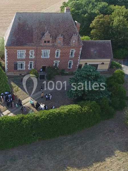 Vue aérienne d'une maison en briques rouges avec un jardin, entourée de haies et de champs, des personnes rassemblées devant.