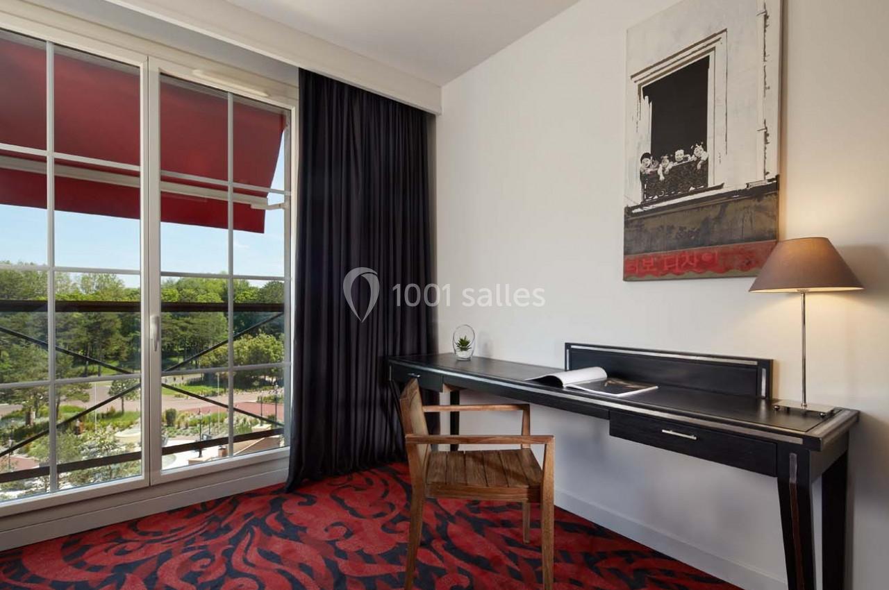 Bureau en bois sombre avec chaise, près d'une fenêtre donnant sur un paysage verdoyant, dans une pièce au sol rouge.