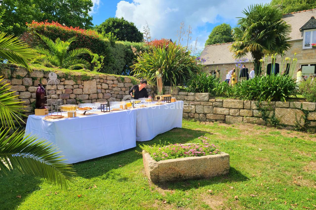 Table dressée en extérieur avec boissons et apéritifs, entourée de verdure et de murs en pierre dans un jardin.
