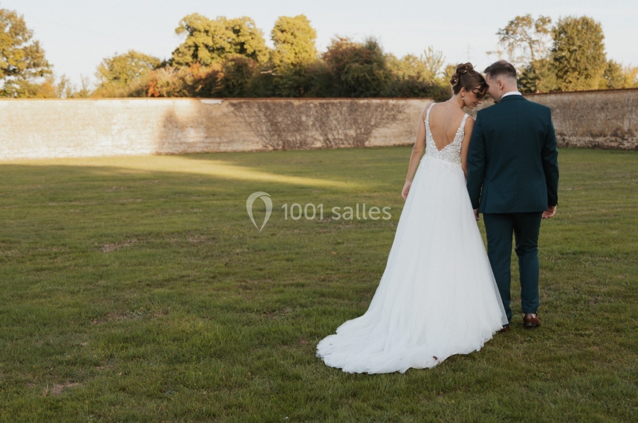 Une mariée en robe blanche et un marié en costume vert marchent côte à côte dans un jardin.