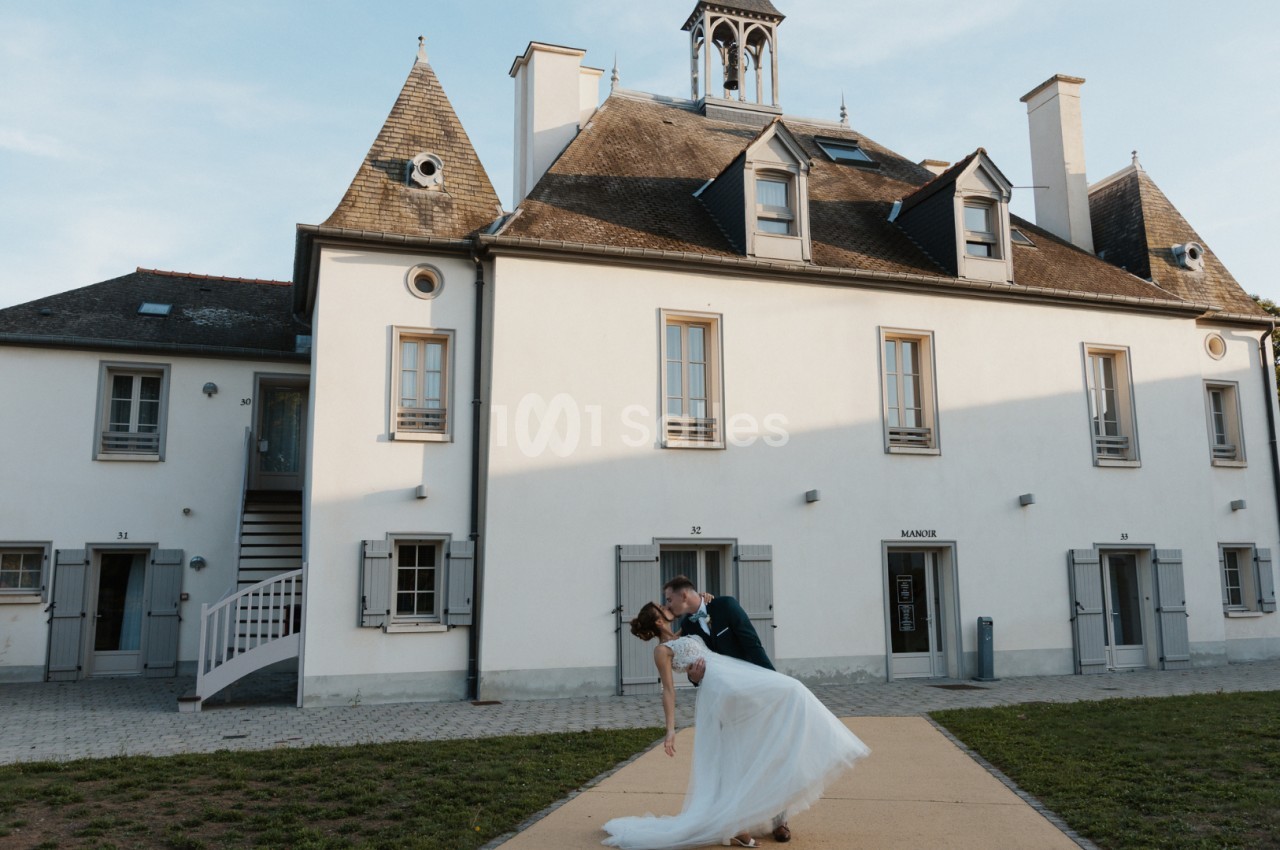 Un couple en tenue de mariage danse devant un bâtiment ancien avec des toits en ardoise et une petite cloche.