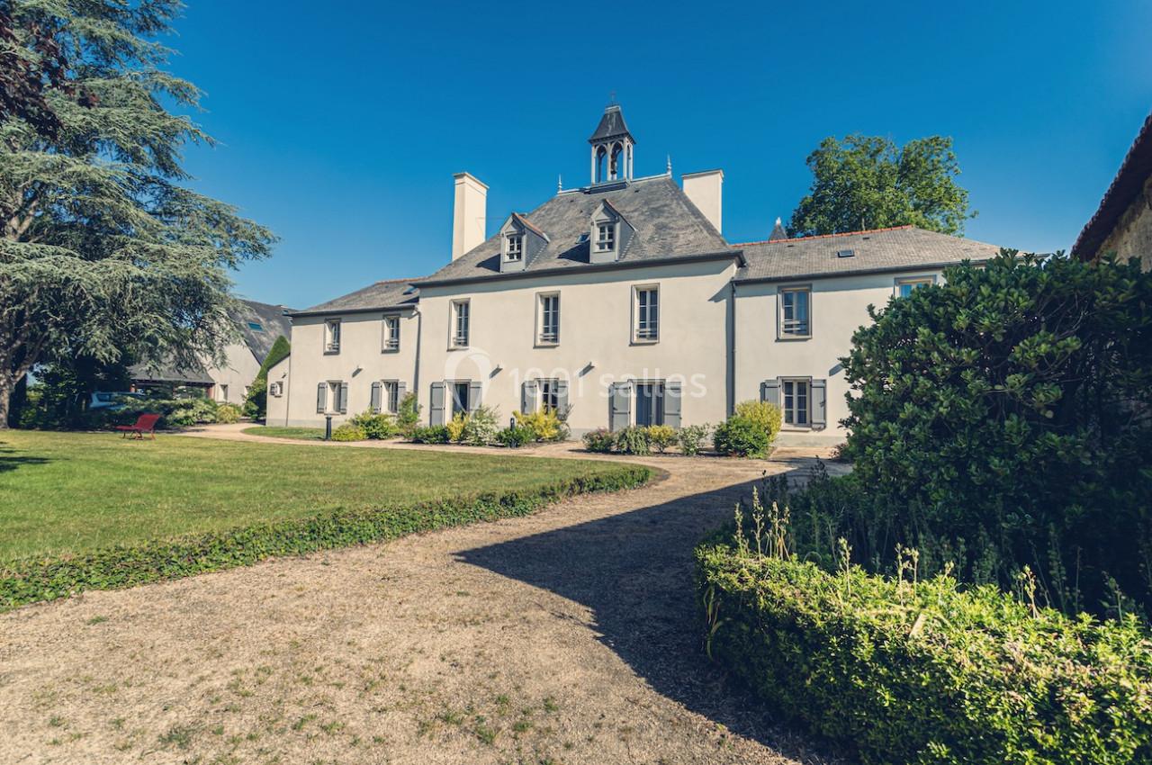 Grande maison ancienne avec façade blanche, toit en ardoise, jardin entretenu et allée en gravier sous un ciel bleu.
