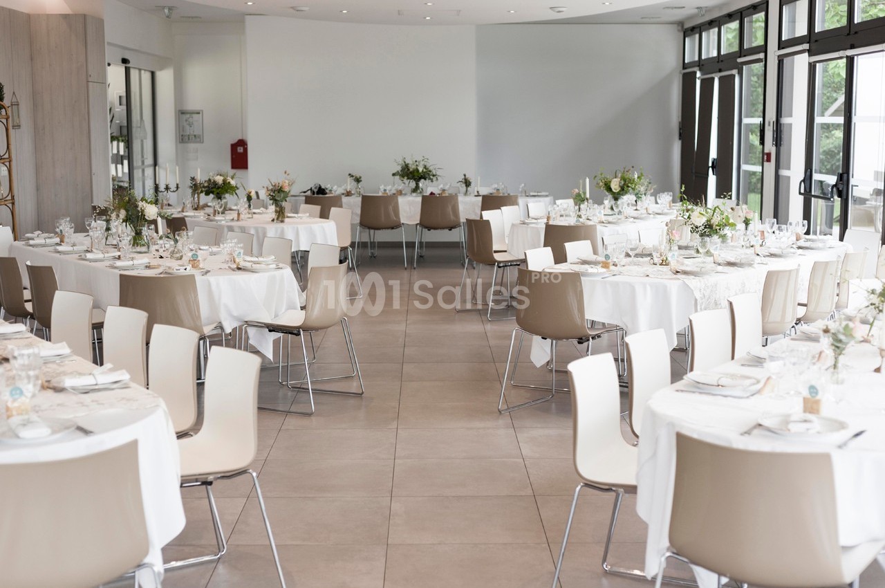 Salle lumineuse aménagée pour un événement, avec des tables rondes dressées et décorées de fleurs.