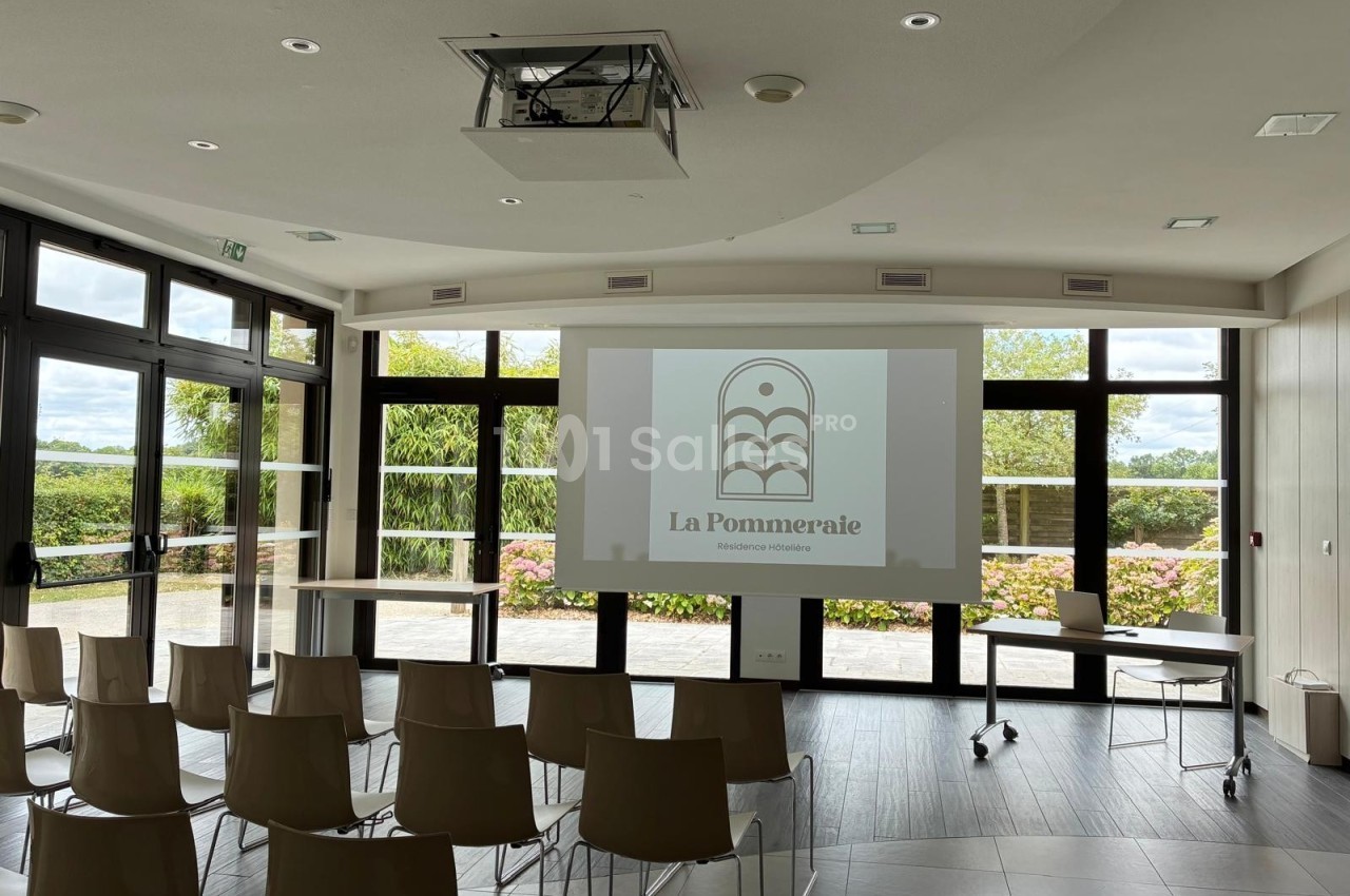 Salle de conférence lumineuse avec chaises alignées, écran de projection affichant un logo et vue sur un jardin extérieur.