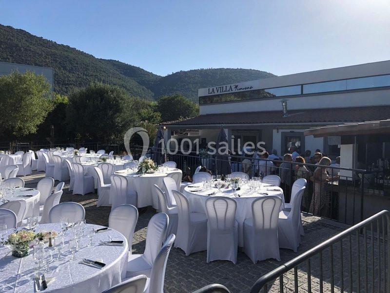 Terrasse extérieure d'un restaurant avec des tables rondes dressées pour un événement, entourée de collines verdoyantes.