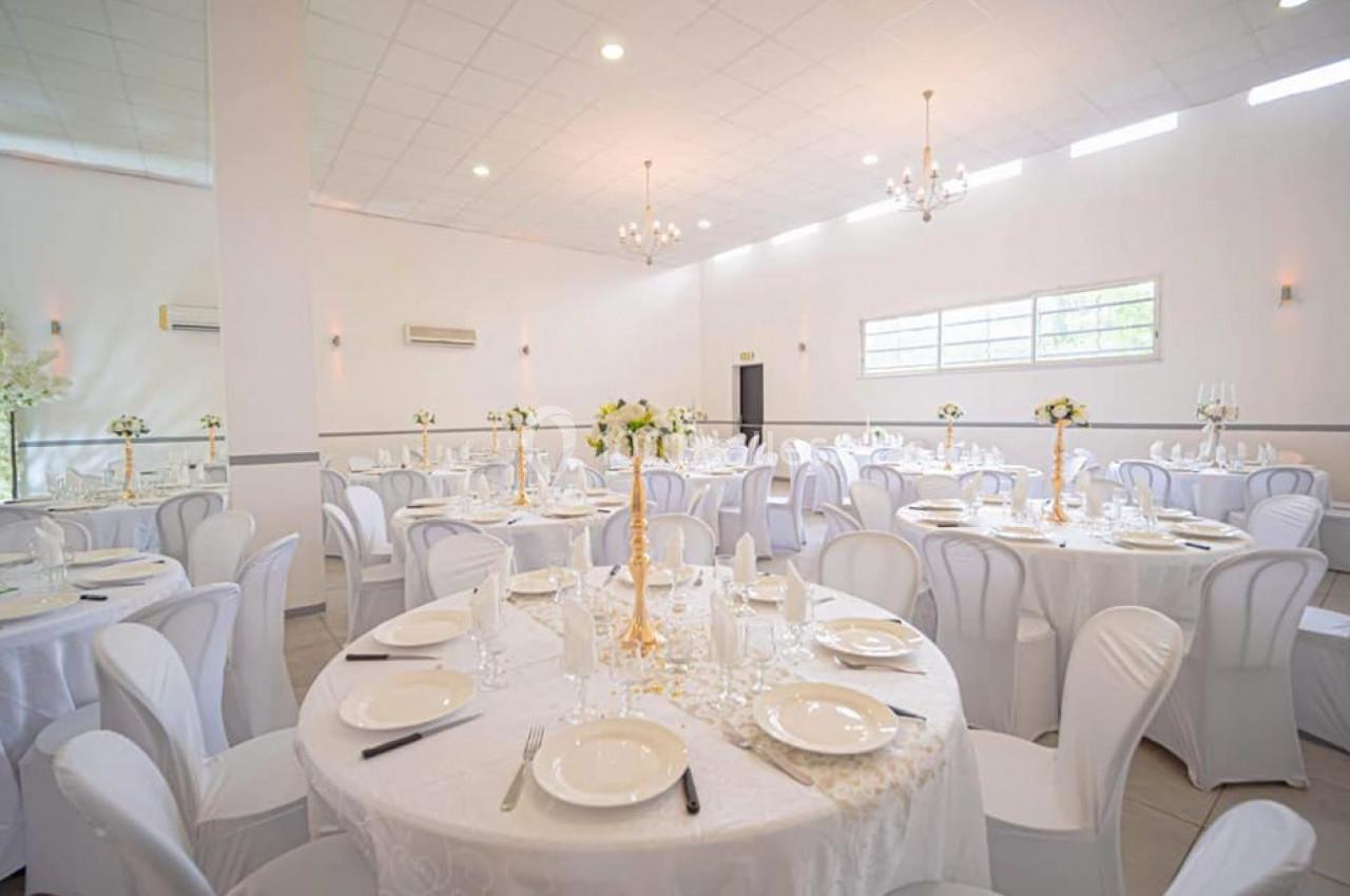 Salle de réception lumineuse avec tables rondes décorées de nappes blanches, vaisselle et centres de table floraux.
