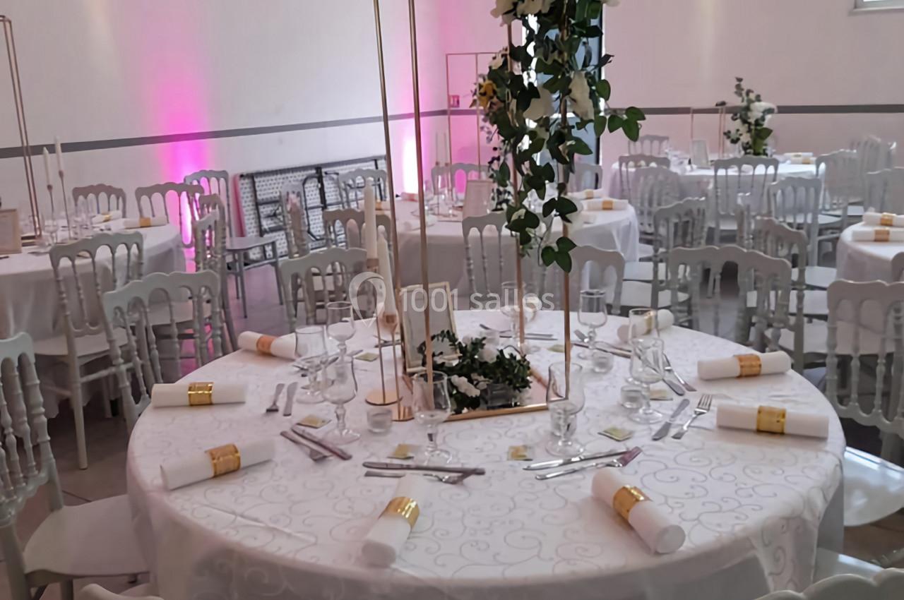 Salle de réception décorée avec des tables rondes, nappes blanches, chaises élégantes et centres de table fleuris.