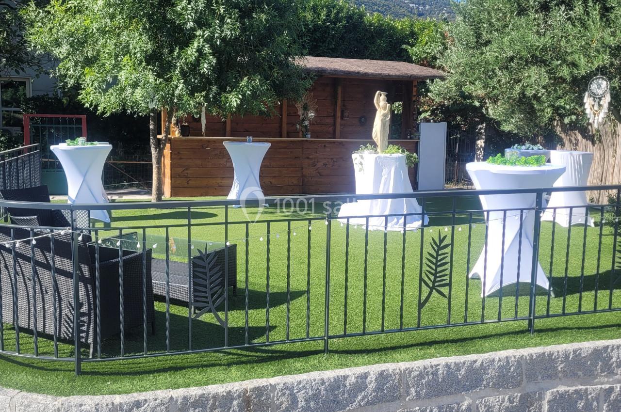Espace extérieur aménagé avec pelouse artificielle, tables hautes, fauteuils en rotin et cabanon en bois, entouré de verdure.