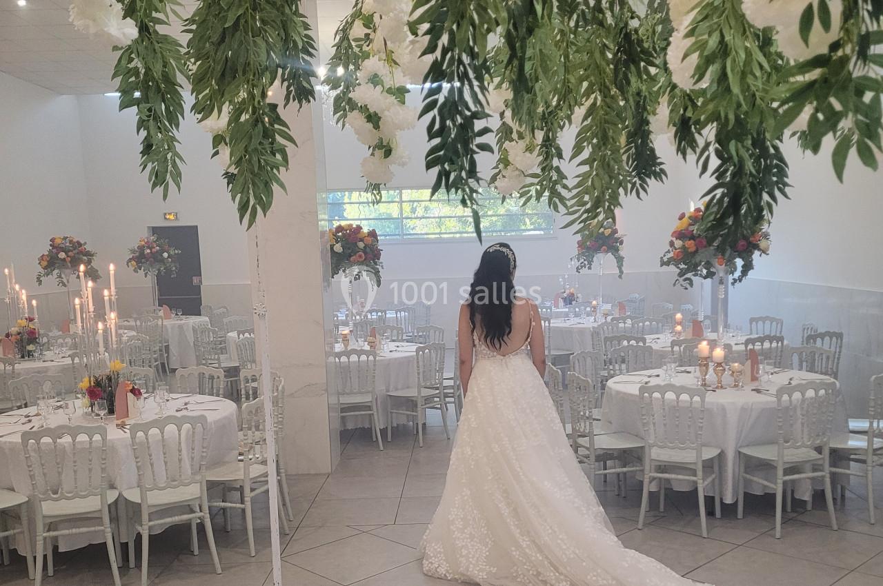 Une mariée en robe blanche regarde une salle de réception décorée de fleurs suspendues et de tables dressées.