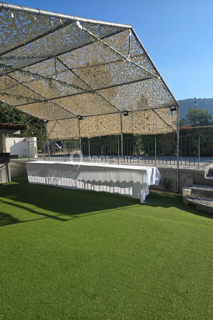 Table drapée de blanc sous une structure ombragée, installée sur une pelouse synthétique par temps ensoleillé.