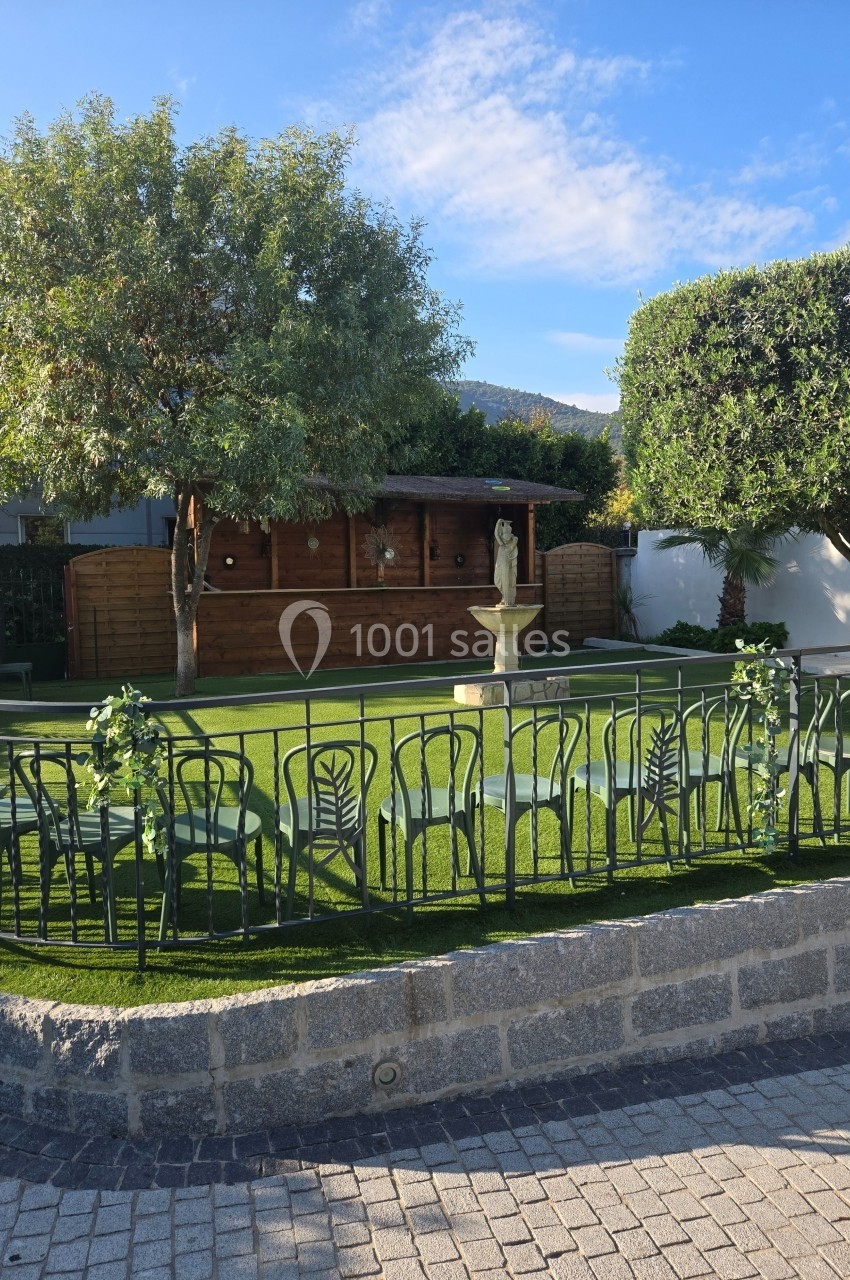 Espace extérieur avec pelouse, fontaine centrale, chaises en métal et cabanon en bois entouré d'arbres.