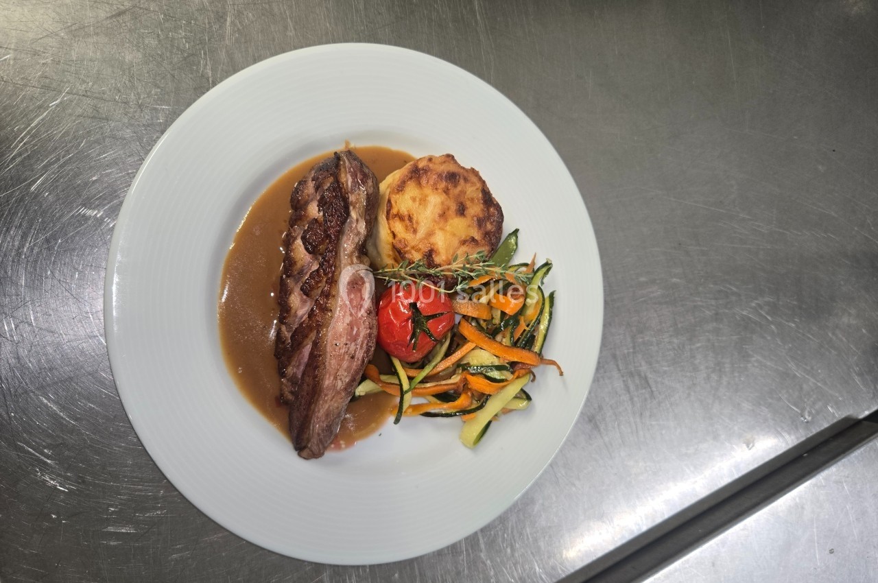 Assiette contenant une pièce de viande avec sauce, gratin de pommes de terre et légumes variés sur une table en inox.