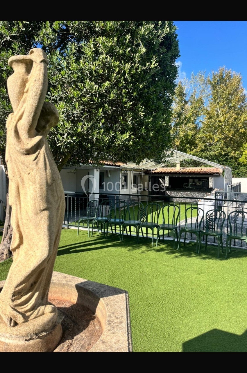 Statue en pierre dans un jardin avec pelouse artificielle, entourée de tables et chaises en métal sous des arbres.