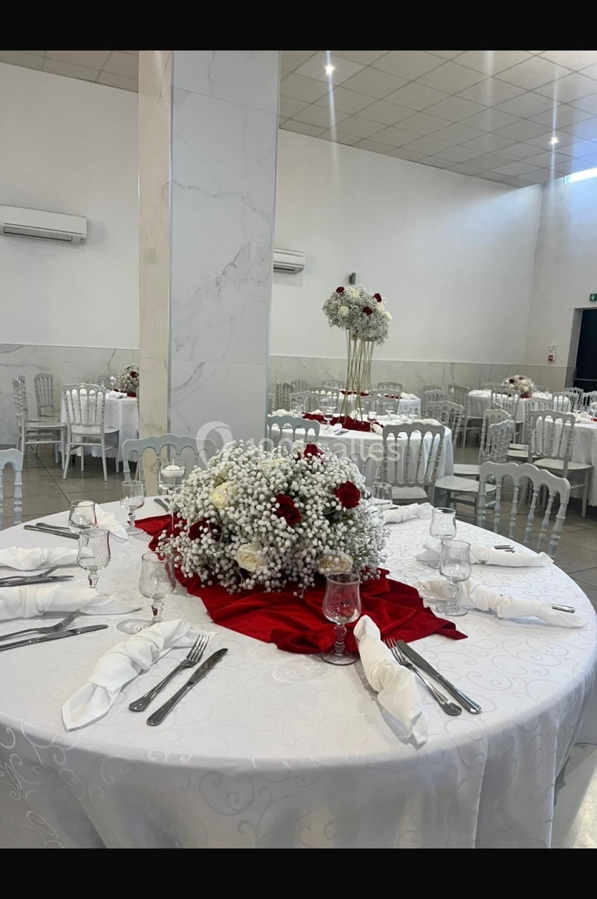 Salle de réception avec tables rondes dressées, nappes blanches, décorations florales rouges et blanches.