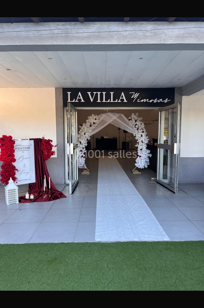 Entrée d'une salle de réception avec arche florale blanche, tapis blanc et décorations rouges sur les côtés.