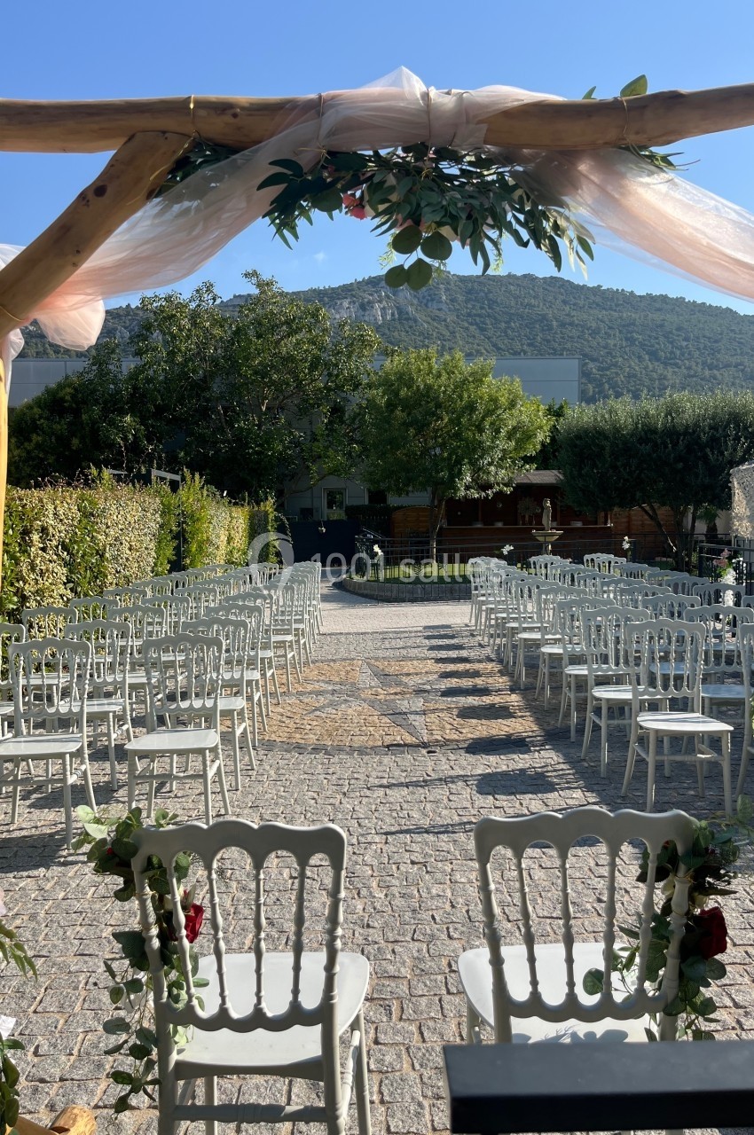 Allée centrale bordée de chaises blanches, vue depuis une arche décorée, dans un jardin ensoleillé avec colline en arrière…