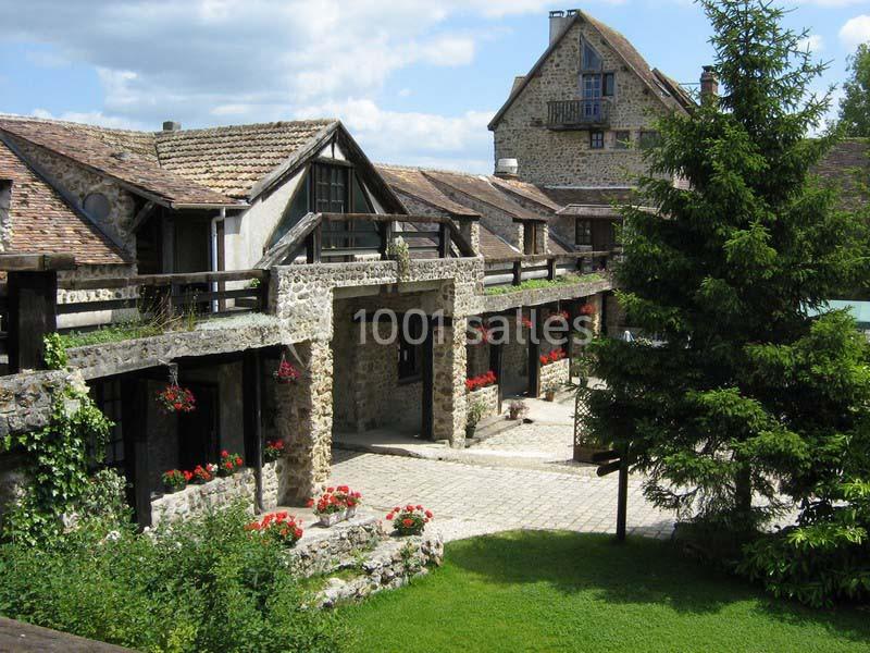Cour pavée bordée de bâtiments en pierre avec balustrades en bois, fleurs rouges et un arbre au premier plan.