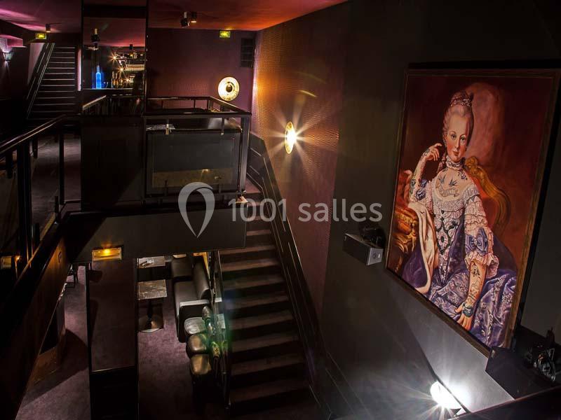 Intérieur d'un espace élégant avec escalier, éclairage tamisé et grand tableau représentant une femme en tenue d'époque.