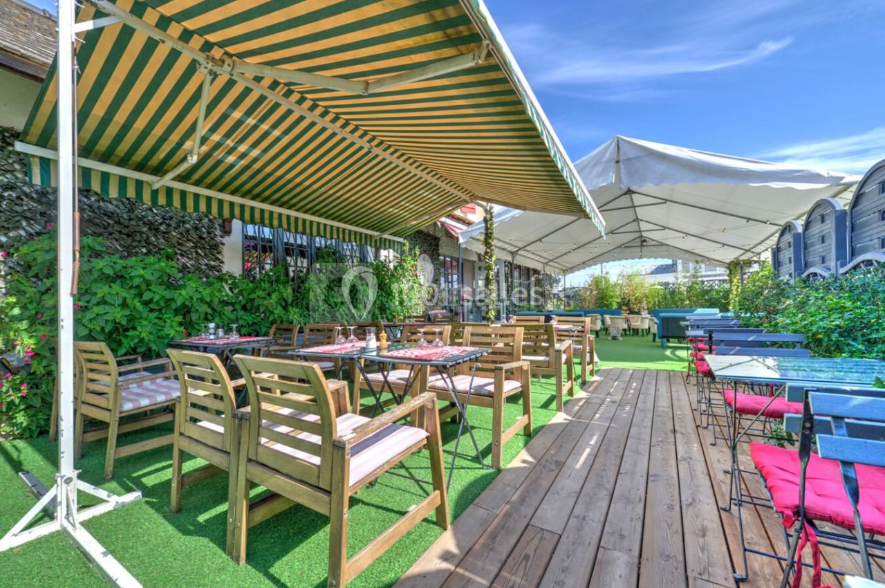 Terrasse extérieure avec tables et chaises en bois sous des auvents, entourée de verdure et de ciel dégagé.