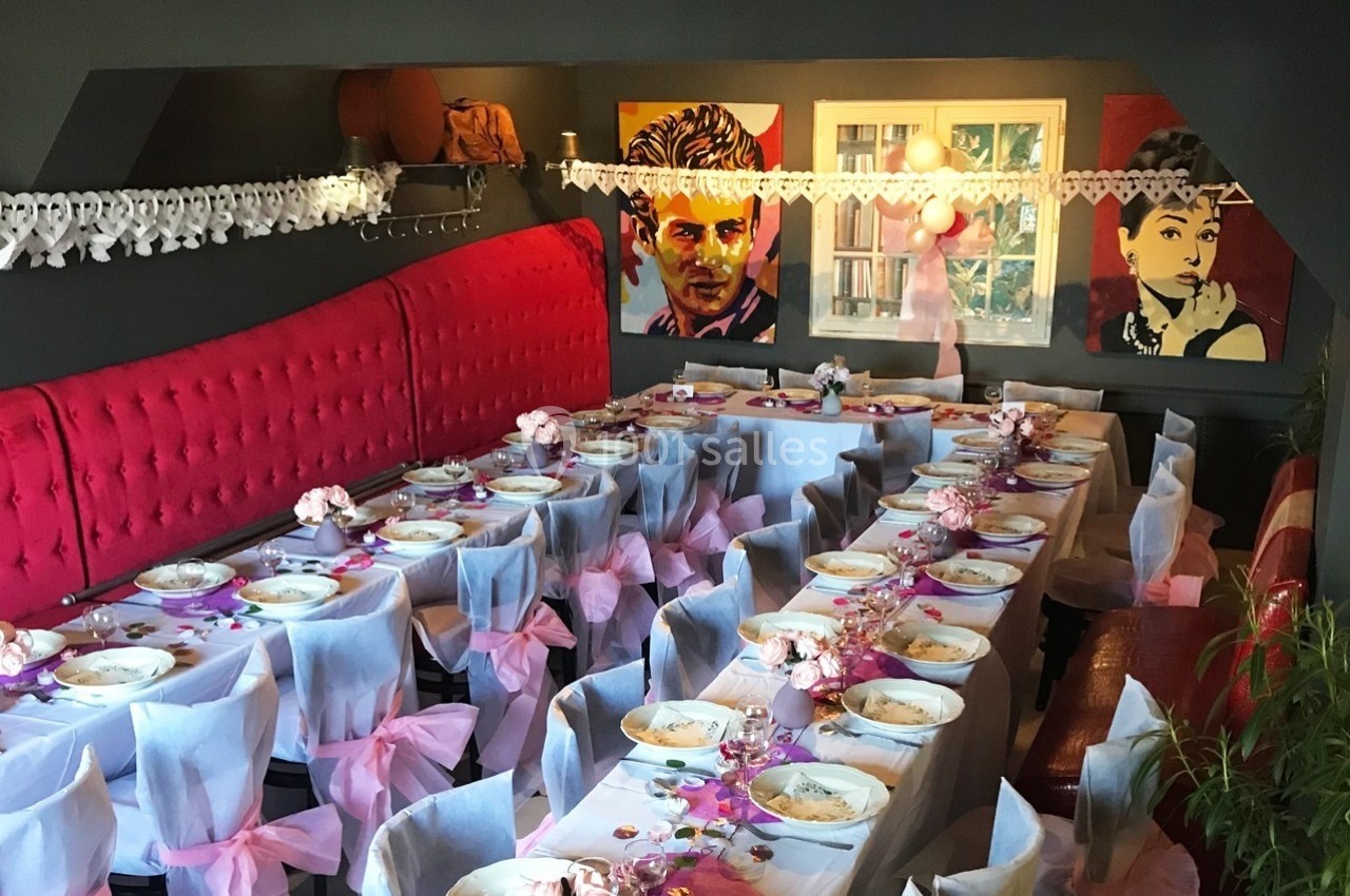 Salle décorée pour un événement, avec tables alignées, nappes blanches, nœuds roses et portraits accrochés au mur.