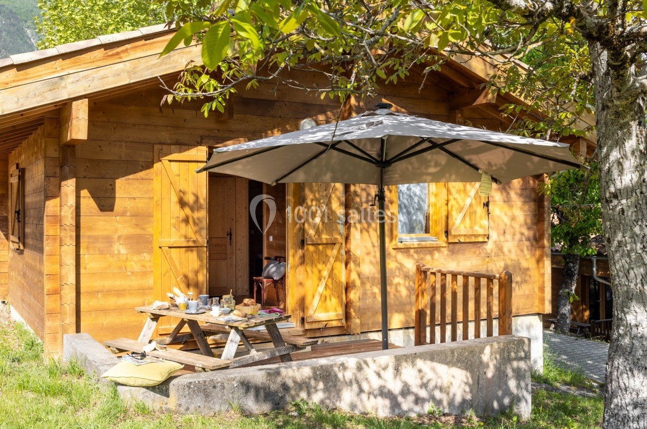 Chalet en bois avec terrasse, table de pique-nique, parasol et arbres environnants par une journée ensoleillée.