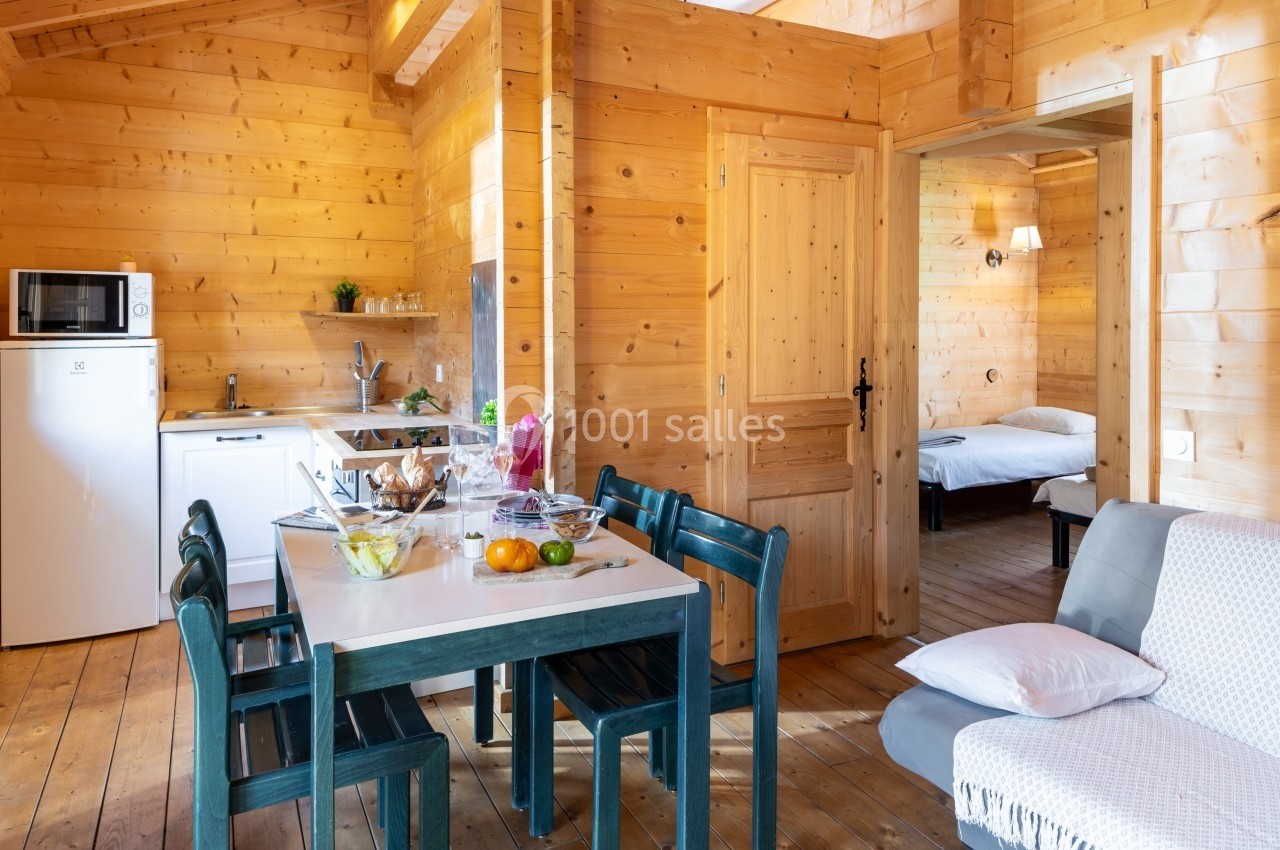 Intérieur d'un chalet en bois avec cuisine équipée, table à manger, salon et vue sur une chambre avec lit.
