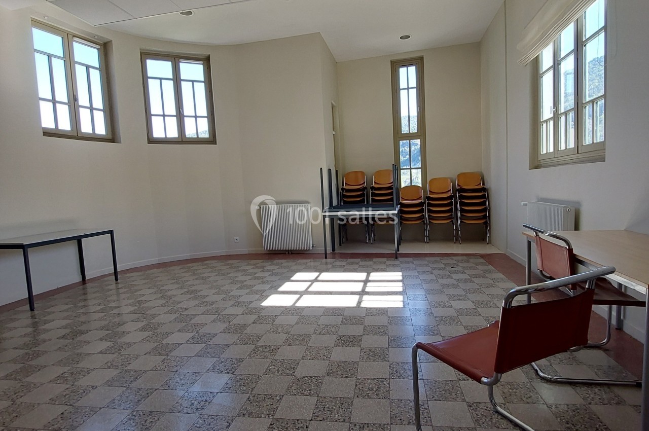 Salle lumineuse avec carrelage en damier, chaises empilées près d'une fenêtre et quelques tables disposées autour.