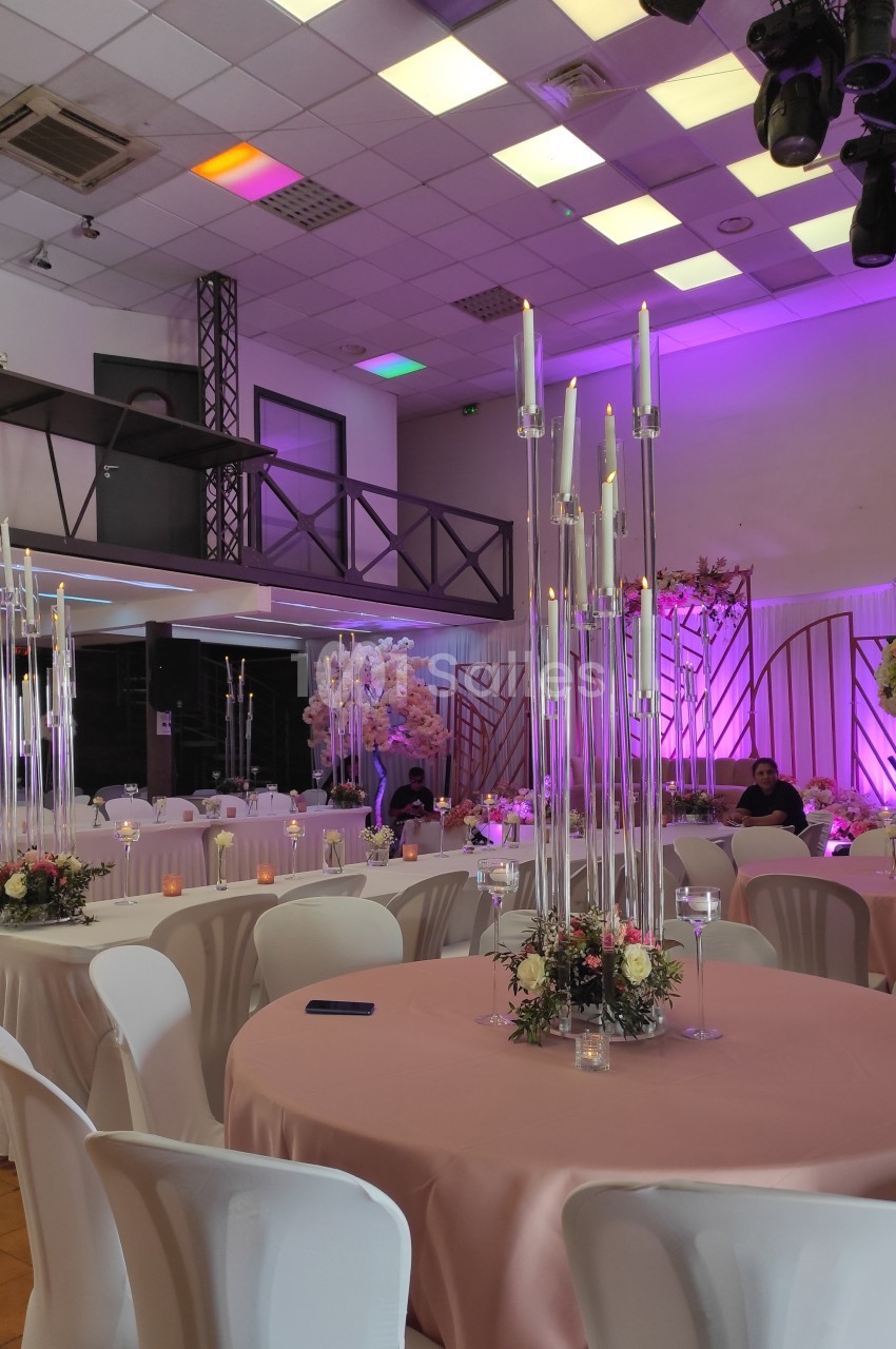 Salle de réception décorée avec des tables rondes, nappes roses, chandeliers hauts et éclairage coloré.