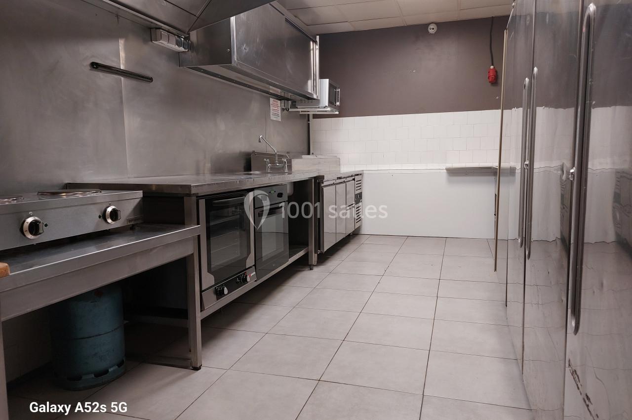Cuisine professionnelle équipée avec plans de travail en inox, four, plaques de cuisson et carrelage au sol.