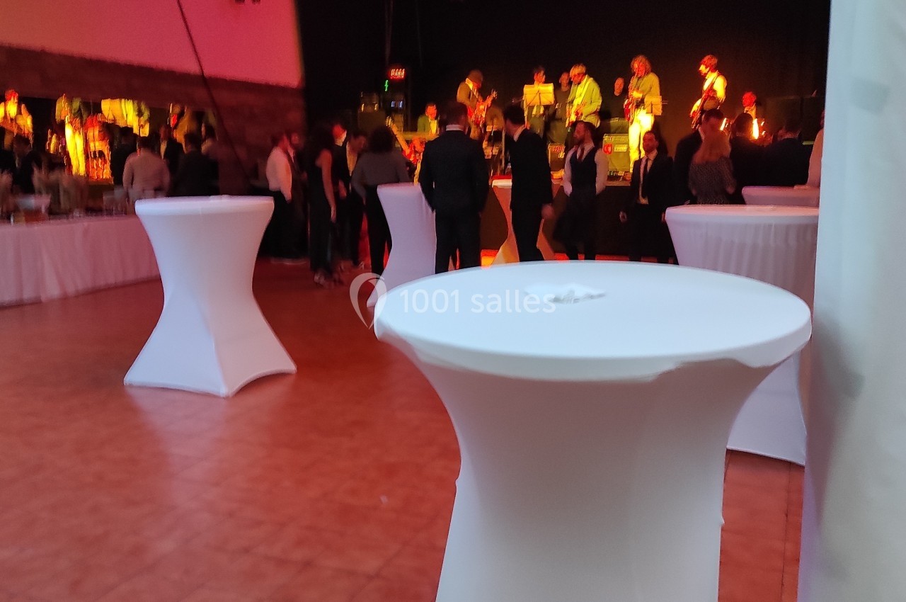 Salle de réception avec tables hautes blanches, invités debout et scène en arrière-plan avec musiciens.