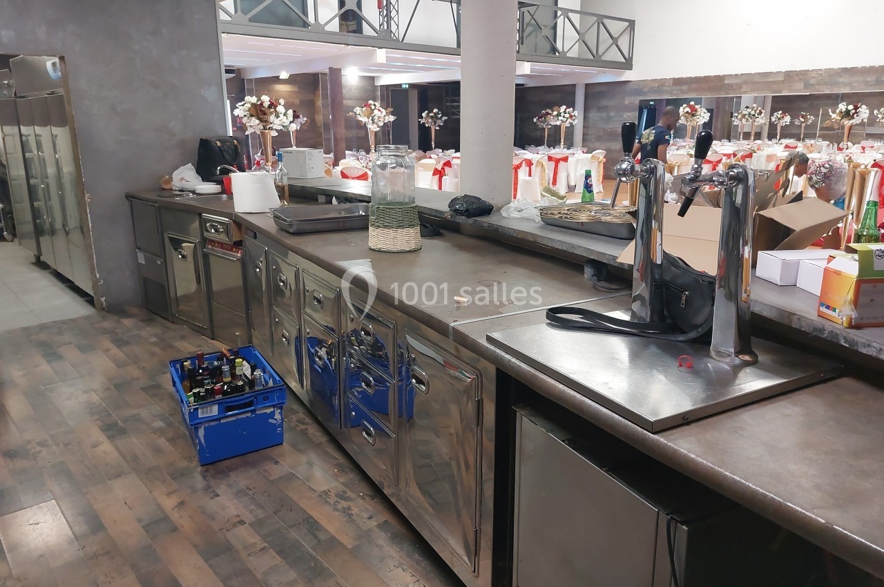 Comptoir de cuisine professionnelle avec équipements, caisses de bouteilles et salle de réception en arrière-plan.