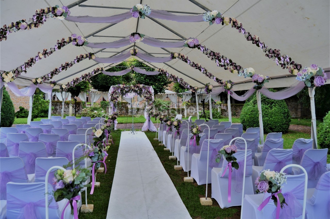 Allée centrale décorée de fleurs violettes et blanches sous une tente pour une cérémonie en plein air.