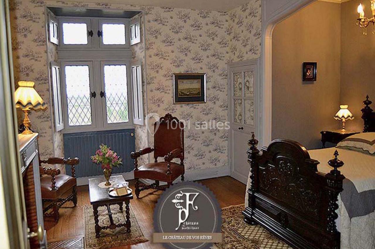 Chambre ancienne avec mobilier en bois sculpté, cheminée, deux fauteuils et fenêtre à vitraux donnant sur l'extérieur.