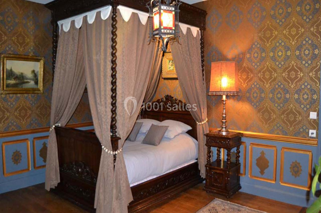Lit à baldaquin en bois sculpté avec rideaux, dans une chambre décorée de motifs classiques et éclairée par une lampe.