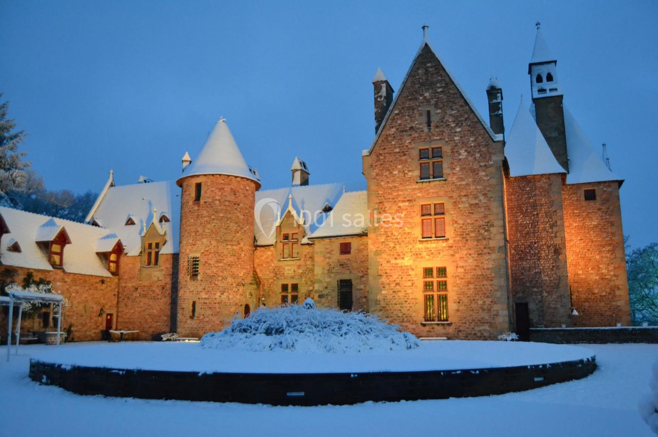 Château en pierre éclairé au crépuscule, entouré de neige et d'arbres sous un ciel hivernal.