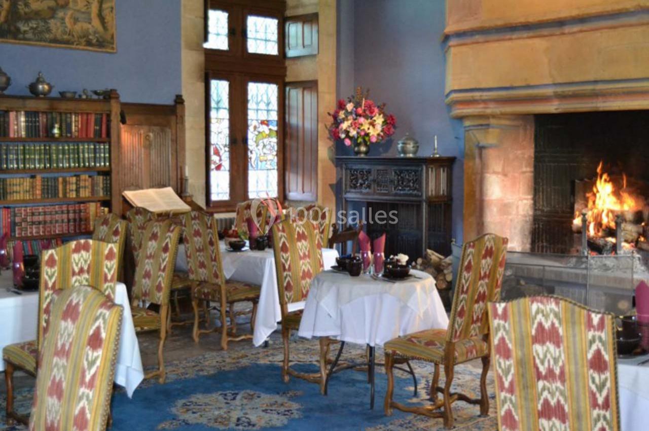 Salle à manger chaleureuse avec cheminée allumée, tables dressées, chaises à motifs et bibliothèque en arrière-plan.