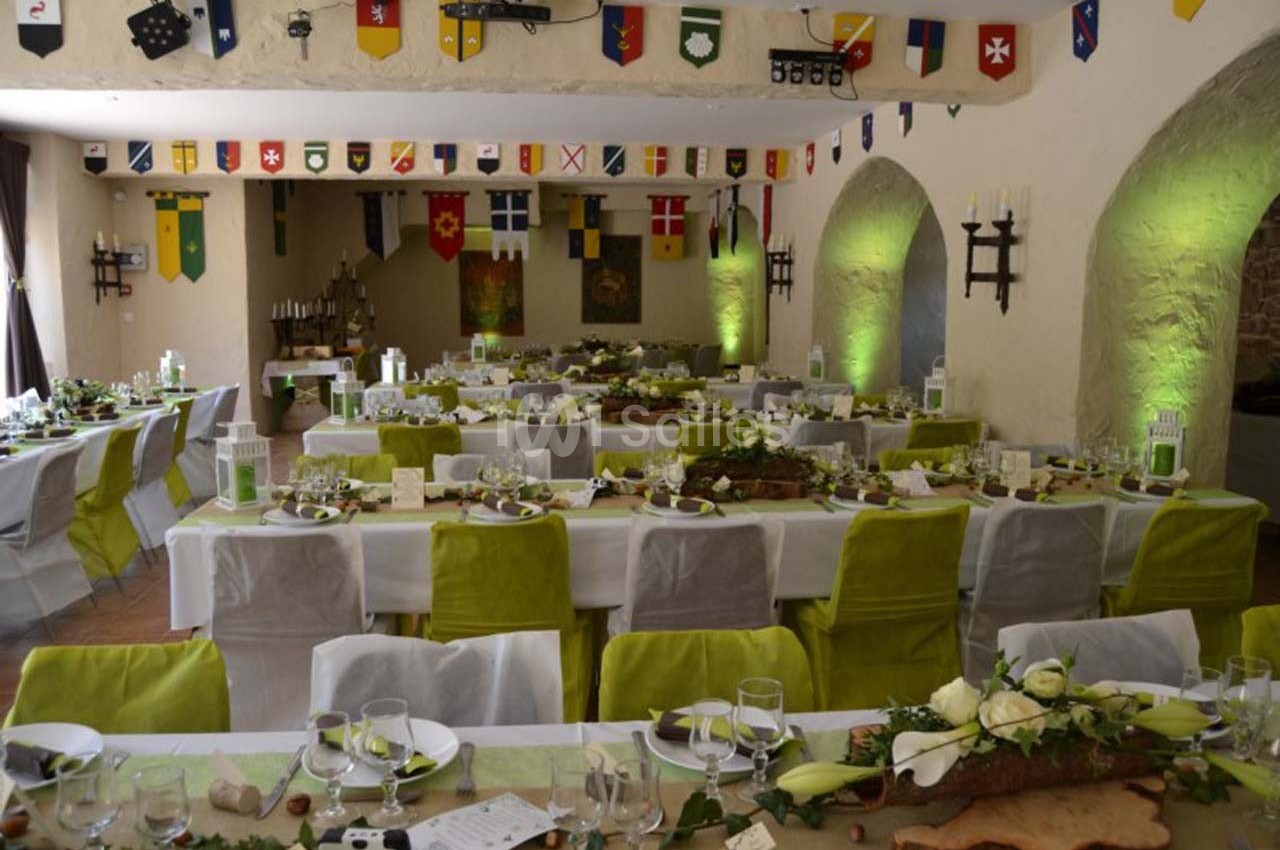 Salle décorée pour un banquet, avec des tables dressées, des chaises vertes et des bannières médiévales suspendues.