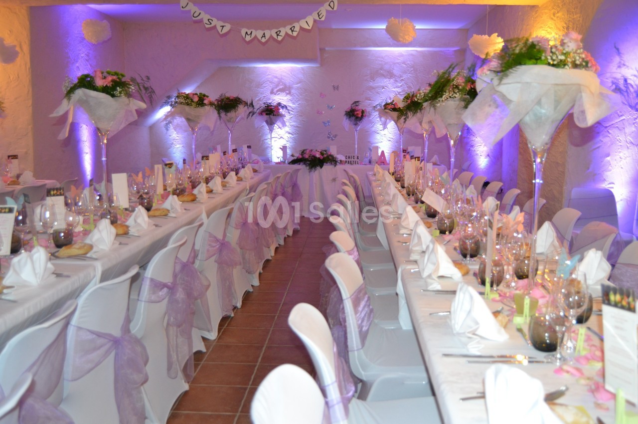 Salle de réception décorée pour un mariage, avec longues tables dressées, nappes blanches et fleurs violettes.