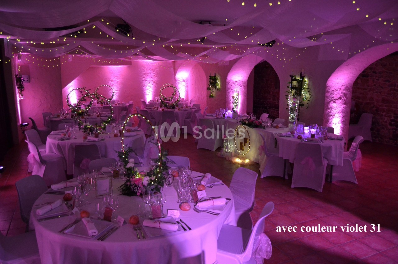 Salle de réception décorée avec des guirlandes lumineuses, des nappes blanches et une ambiance lumineuse violette.